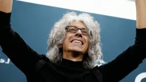 Persona con capelli grigi ricci e occhiali sorride con le braccia sollevate