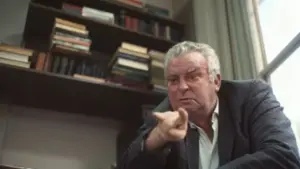 Uomo anziano in un ufficio con una libreria alle spalle, indica con decisione