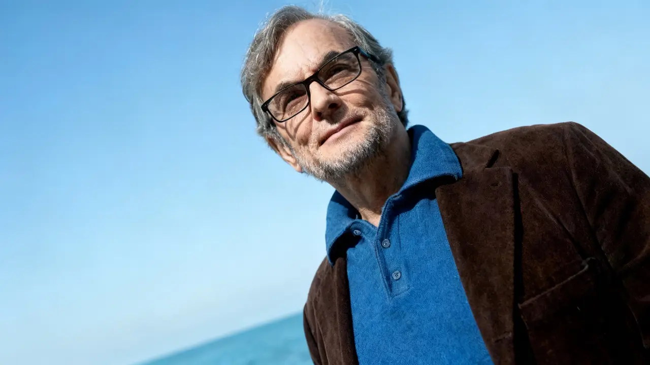 Un uomo con occhiali in posa all'aperto vicino al mare, con cielo sereno sullo sfondo