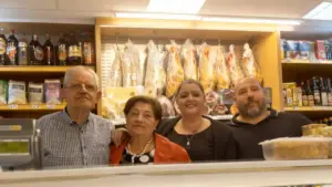 Quattro persone dietro il bancone di un negozio alimentare con salumi appesi e scaffali pieni