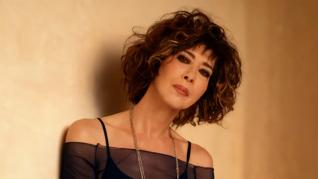 Una donna con capelli ricci e trucco marcato posa contro una parete beige