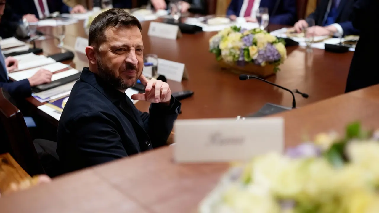 Una persona seduta a un tavolo di conferenza guarda verso la fotocamera, circondata da altri partecipanti.