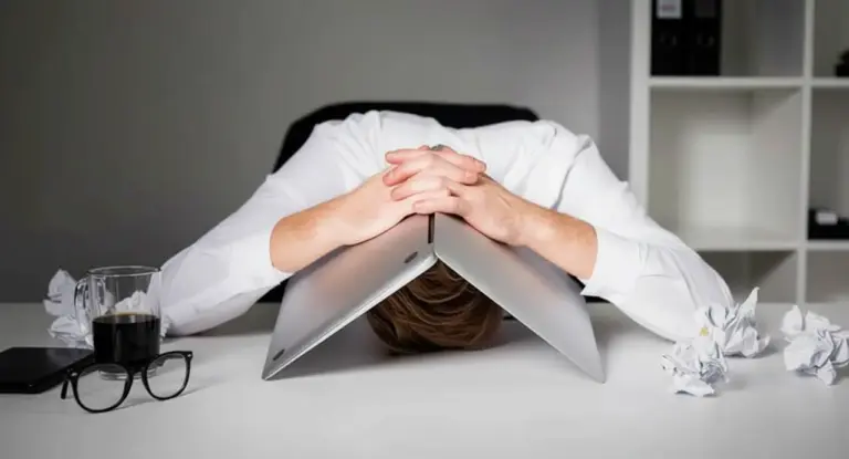 Persona con la testa abbassata su un laptop chiuso, circondata da fogli accartocciati