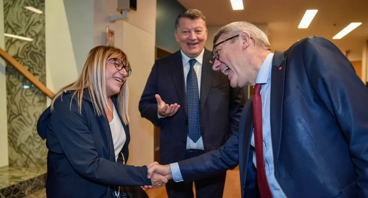 Tre persone in abiti formali si salutano cordialmente in un ambiente interno