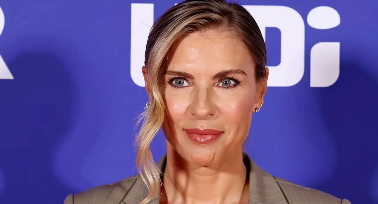 Una donna con capelli biondi pettinati di lato posa davanti a uno sfondo blu