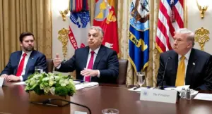 Tre persone sedute a un tavolo durante un incontro ufficiale con bandiere militari sullo sfondo