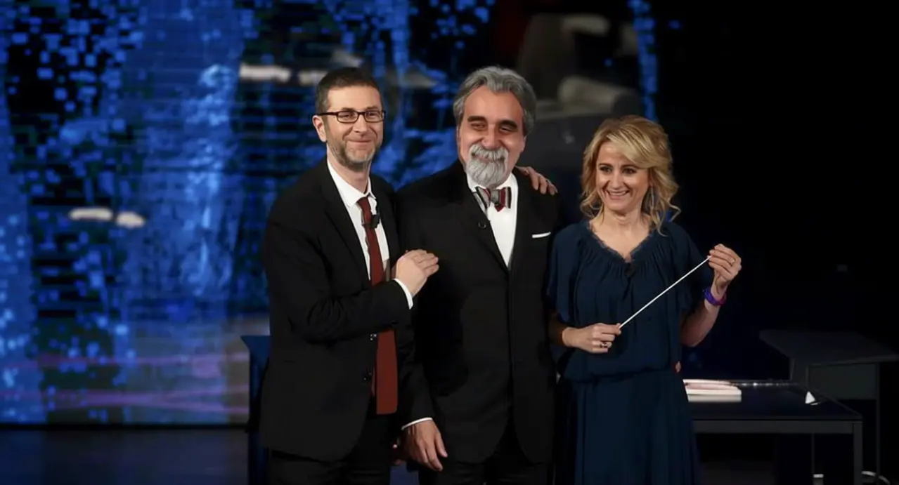 Tre persone sorridenti in abbigliamento elegante su un palco televisivo