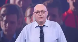 Una persona calva con occhiali e cravatta parla di fronte a un pubblico sfocato