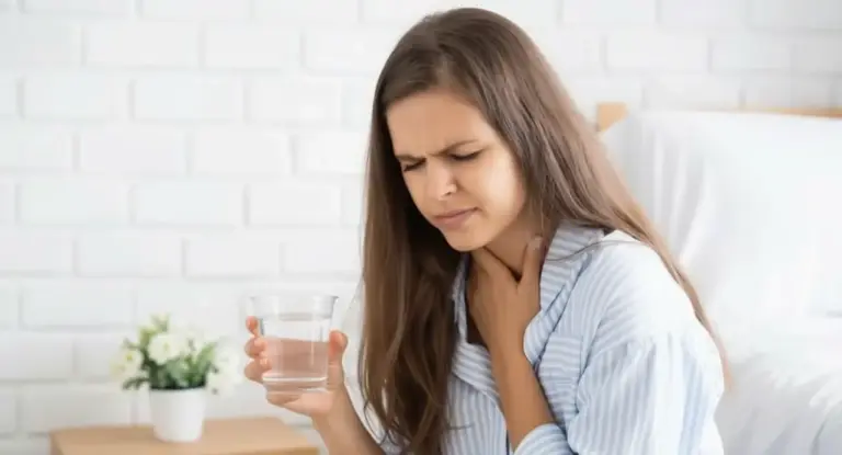 Persona seduta su un letto che tiene la gola con una mano e un bicchiere d'acqua con l'altra, esprimendo disagio